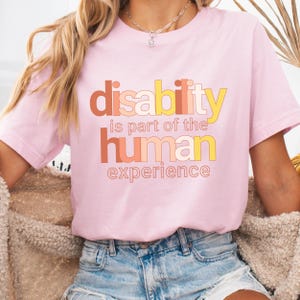 May include: A pink t-shirt with the text "disability is part of the human experience" in colorful, layered letters.
