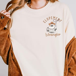 Puede incluir: Sudadera de color crema con un gráfico de un fantasma con un sombrero de vaquero y un lazo. El texto "Elopement Wrangler" está impreso encima y debajo de la ilustración del fantasma en color marrón. La modelo lleva una chaqueta de pana marrón.