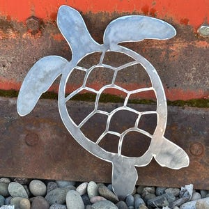 May include: A metal cutout of a sea turtle with a detailed shell design. The turtle is painted silver and is sitting on a bed of small, gray rocks.