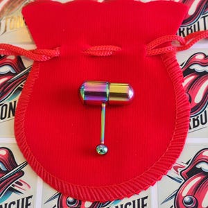 Pode incluir: Uma barra de língua arco-íris com uma bola em forma de pílula em uma extremidade e uma bola menor na outra extremidade. A barra é exibida em uma bolsa de veludo vermelho.