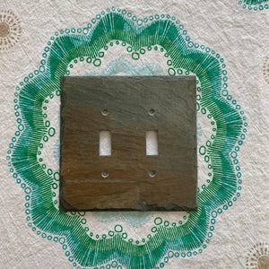 May include: A square, dark gray slate double light switch cover. The cover has two rectangular openings for the switches and two small holes for mounting. The slate has a natural, textured surface. The background is a white fabric with a green and gold pattern.