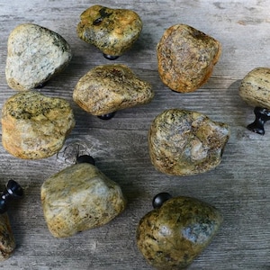 May include: A collection of natural stone cabinet knobs. The knobs are various shapes and sizes, with a mix of tan, brown, and gray tones. Each stone is mounted on a black metal base, creating a unique and rustic look for home decor.