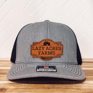 Op de afbeelding: Een grijze en zwarte trucker-pet met een bruin leren patch waarop "Lazy Acres Farms" staat met een tractorsilhouet. De patch heeft decoratieve stiksels rond de randen. De pet heeft een zwart mesh-achterpaneel en een verstelbare Richardson 112-riem.