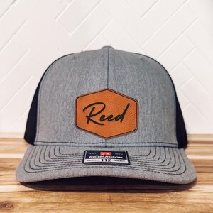 Könnte beinhalten: Eine graue und schwarze Trucker-Kappe mit einem braunen Leder-Patch, auf dem "Reed" in Kursivschrift steht. Die Kappe ist verstellbar und hat ein schwarzes Netzrücken.