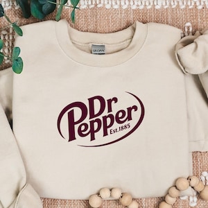 May include: A beige sweatshirt with the Dr Pepper logo in maroon. The logo is in a stylized script within an oval, with "Est. 1885" below. The sweatshirt is on a patterned surface with decorative beads.