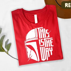 A red t-shirt with a white graphic design and text printed on it, along with a brown hat and some greenery.