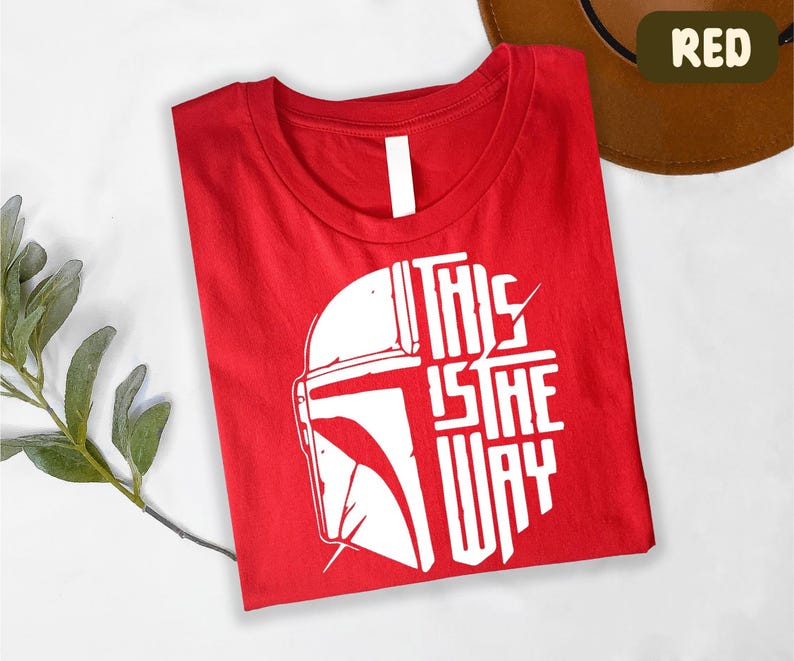 A red t-shirt with a white graphic design and text printed on it, along with a brown hat and some greenery.