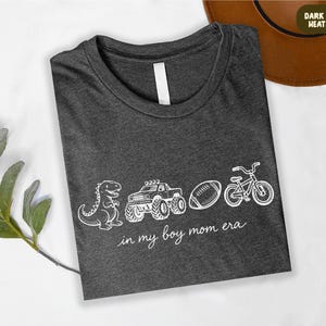 A gray t-shirt with white text and illustrations of a dinosaur, truck, bicycle, and football, along with the phrase "in my boy mom era" printed on it.