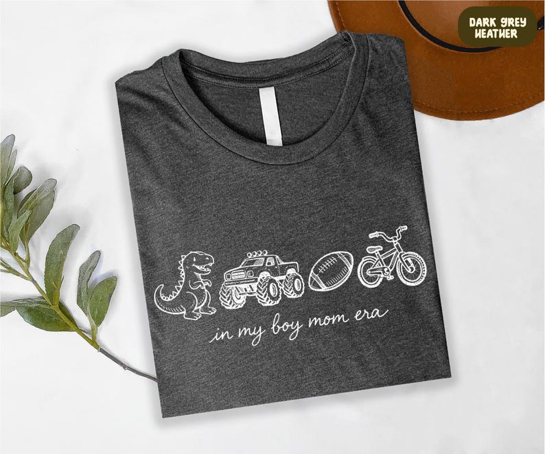 A gray t-shirt with white text and illustrations of a dinosaur, truck, bicycle, and football, along with the phrase "in my boy mom era" printed on it.
