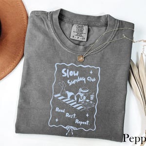 May include: A gray t-shirt with the text "Slow Sunday Club" and an illustration of a person relaxing on a beach towel. The shirt has a crew neck and short sleeves. A necklace with a pendant is on the shirt.