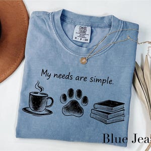 May include: Blue jean colored t-shirt with the phrase "My needs are simple" and illustrations of a coffee cup, paw print, and books. A gold necklace and a brown hat are also visible.