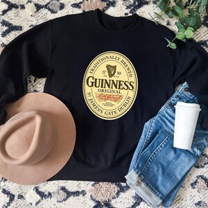 a black sweatshirt with a Guinness logo, a tan hat, a pair of blue jeans, and a white coffee cup, all laid out on a patterned rug.