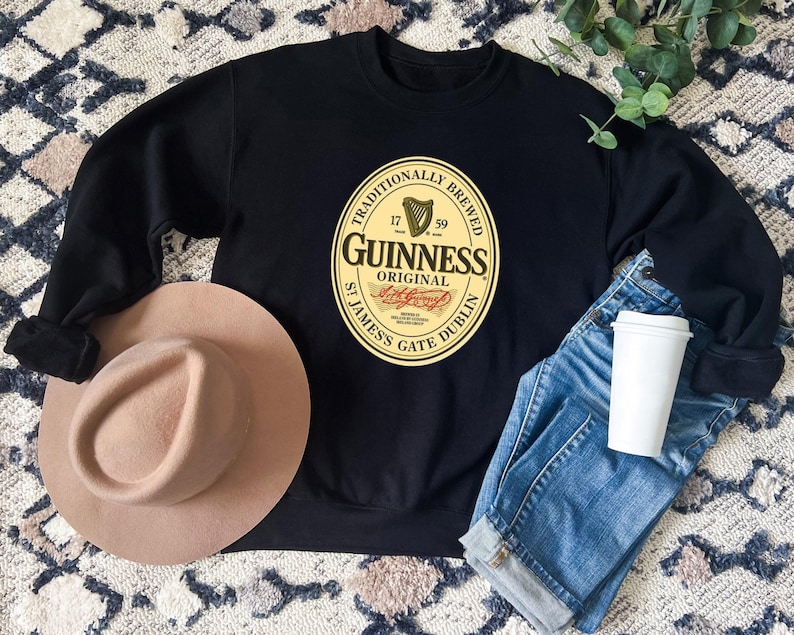 a black sweatshirt with a Guinness logo, a tan hat, a pair of blue jeans, and a white coffee cup, all laid out on a patterned rug.