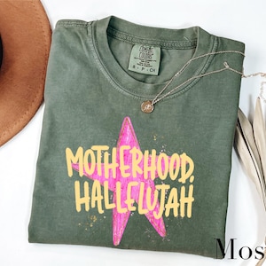 May include: A moss green t-shirt with the words "MOTHERHOOD, HALLELUJAH" in yellow text over a pink star. A gold necklace with a pendant is draped on the shirt. The shirt is a Comfort Colors brand.