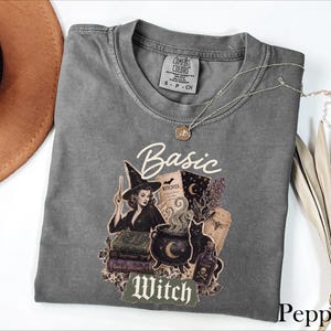 May include: A charcoal gray short-sleeve t-shirt featuring a graphic print of a witch, a cauldron, books, and a black cat. The text reads Basic Witch. The shirt has a crew neckline and a Comfort Colors brand label on the inside collar area.
