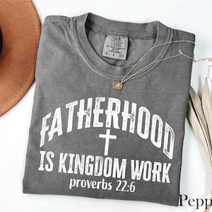 May include: A gray, short-sleeved t-shirt with the text "FATHERHOOD IS KINGDOM WORK" and "proverbs 22:6" printed in white. A silver necklace with a pendant is draped across the shirt. A brown hat and dried leaves are in the background.