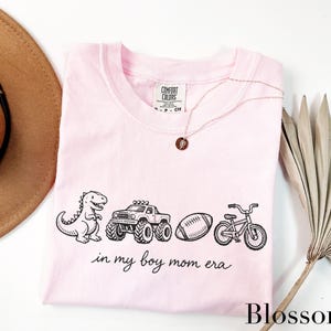 A pink t-shirt with a graphic design of a dinosaur, truck, bicycle, and football, along with the text "in my boy mom era" printed on it. The t-shirt is laid out on a white surface, with a brown hat, a necklace, and a palm frond visible in the background.