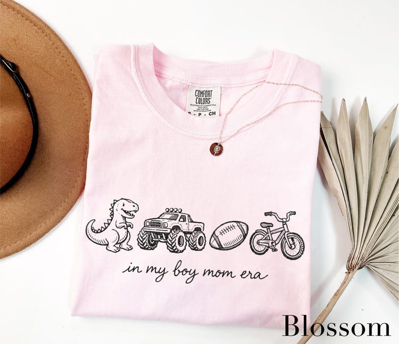 A pink t-shirt with a graphic design of a dinosaur, truck, bicycle, and football, along with the text "in my boy mom era" printed on it. The t-shirt is laid out on a white surface, with a brown hat, a necklace, and a palm frond visible in the background.
