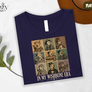 a navy blue t-shirt with a graphic design of nine dog illustrations arranged in a 3x3 grid, each depicting a different dog character. The text "IN MY WISHBONE ERA" is printed below the image.