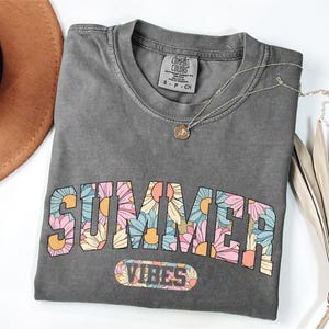 a gray t-shirt with a floral design and the text "SUMMER VIBES" printed on it. The t-shirt is placed on a white surface, along with a brown hat, a necklace, and some dried leaves.