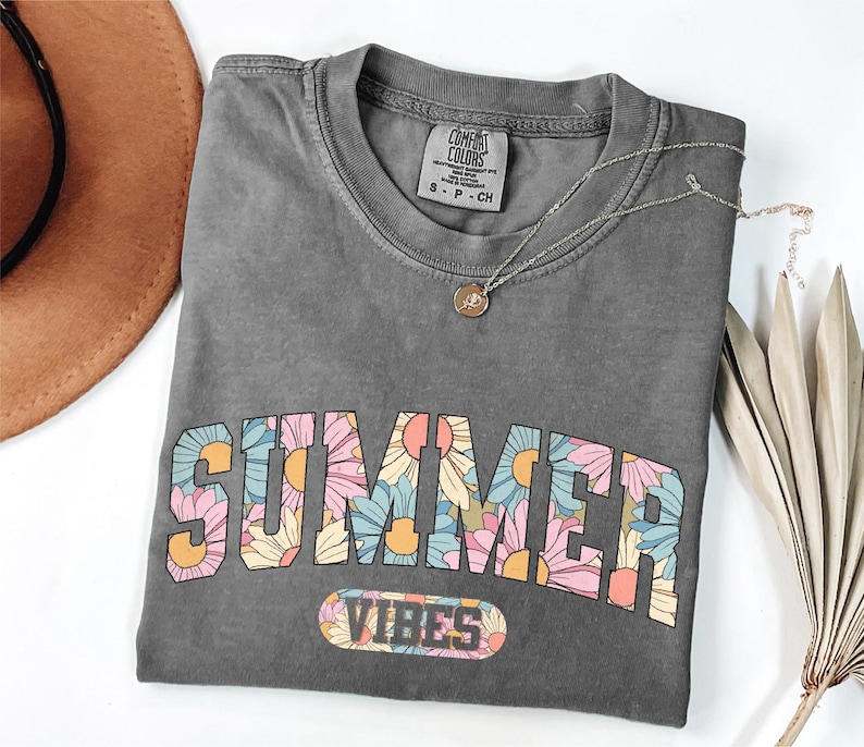 a gray t-shirt with a floral design and the text "SUMMER VIBES" printed on it. The t-shirt is placed on a white surface, along with a brown hat, a necklace, and some dried leaves.