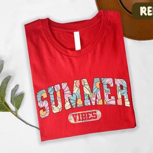 A red t-shirt with the word "SUMMER" printed on it, along with the word "VIBES" below it. The t-shirt is folded and placed on a white surface, with a brown hat visible in the background.