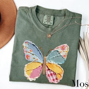 May include: Moss green t-shirt with a colorful butterfly applique. The butterfly wings are patchwork with floral, gingham, and solid fabric pieces. A gold necklace with a pendant rests on the shirt. The shirt has a Comfort Colors tag.