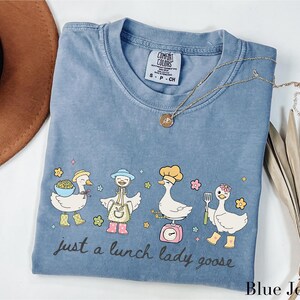May include: Blue jean colored t-shirt with a whimsical illustration of four cartoon geese, each wearing a different chef's hat or apron. The text "just a lunch lady goose" is written in a script font. A gold necklace is draped across the shirt.