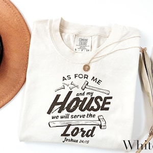 May include: White Comfort Colors t-shirt with the text "AS FOR ME and my HOUSE we will serve the Lord Joshua 24:15" in black. The design includes hammers and nails. A gold necklace and a brown hat are also visible.