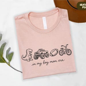 A pink t-shirt with a graphic design of a dinosaur, truck, bicycle, and football, along with the text "in my boy mom era" printed on it.