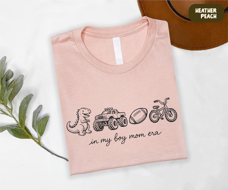 A pink t-shirt with a graphic design of a dinosaur, truck, bicycle, and football, along with the text "in my boy mom era" printed on it.
