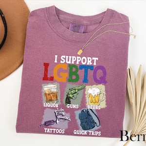 May include: A berry-colored short-sleeve t-shirt featuring the white text I SUPPORT LGBTQ in a rainbow-colored font. Below are five illustrations with labels: a glass of liquor, a green revolver labeled GUNS, a beer mug, a tattoo machine, and a backpack.