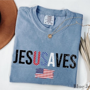 May include: A blue jean-colored t-shirt with the words "JESUS SAVES" in a distressed font. The letters "USA" are filled with a red, white, and blue stars and stripes pattern, and an American flag graphic is below the text.
