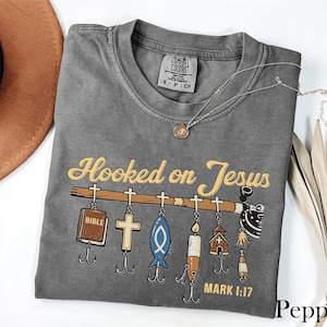 May include: A gray t-shirt with the text "Hooked on Jesus" in yellow script. The design features fishing lures with religious symbols. The shirt has a crew neck and short sleeves. A silver necklace with a pendant is also visible.