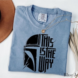 A blue t-shirt with a graphic design of a helmet and the text "THIS IS THE WAY" printed on it, along with a necklace and a brown hat.