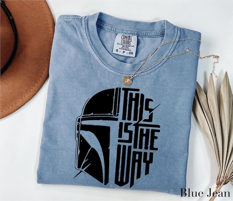 A blue t-shirt with a graphic design of a helmet and the text "THIS IS THE WAY" printed on it, along with a necklace and a brown hat.