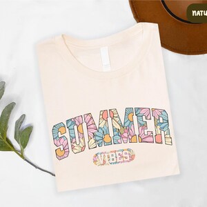 a white t-shirt with a colorful floral design and the word "SUMMER" printed on it, along with a brown hat and some greenery.