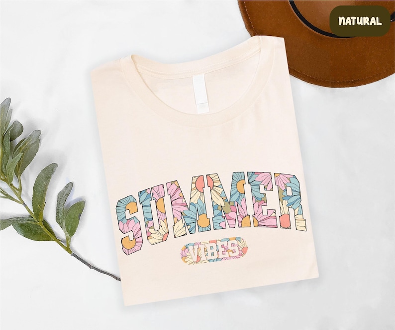 a white t-shirt with a colorful floral design and the word "SUMMER" printed on it, along with a brown hat and some greenery.