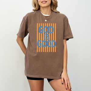 A woman wearing a brown t-shirt with the text "GOD IS GOOD" printed on it.