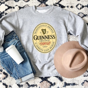 A gray sweatshirt with a Guinness logo, a pair of blue jeans, a beige hat, and a white coffee cup are arranged on a patterned rug.