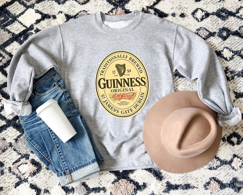 A gray sweatshirt with a Guinness logo, a pair of blue jeans, a beige hat, and a white coffee cup are arranged on a patterned rug.