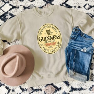 a beige sweatshirt with a Guinness logo, a pink hat, and a pair of blue jeans laid out on a patterned rug.
