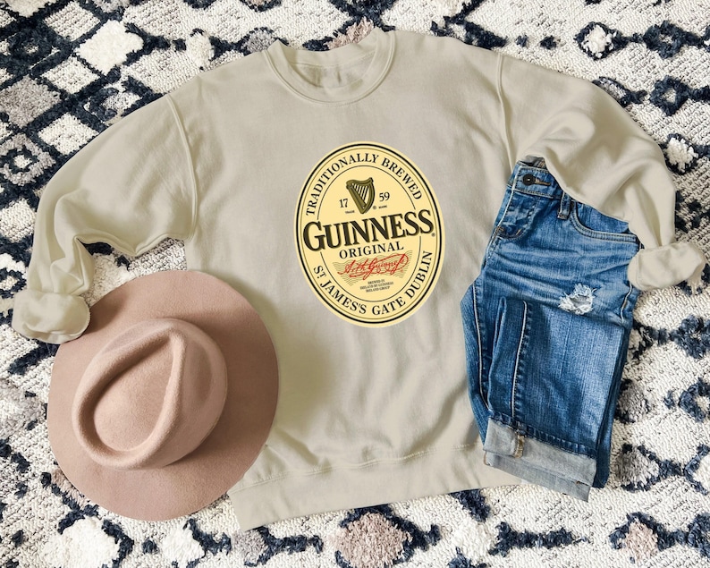 a beige sweatshirt with a Guinness logo, a pink hat, and a pair of blue jeans laid out on a patterned rug.