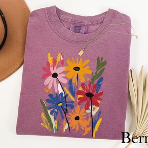 a purple t-shirt with a colorful floral design, a brown hat, and a fan, all arranged on a white background.
