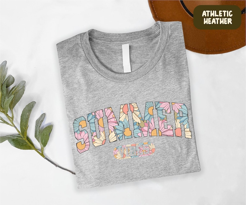 A gray t-shirt with the word "SUMMER" printed on it, along with a brown hat and some greenery, all placed on a white surface.