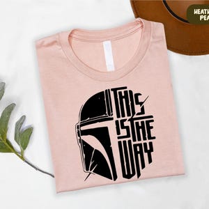 A pink t-shirt with a graphic design featuring a helmet and the text "THIS IS THE WAY" is displayed on a white surface, along with a brown hat and some greenery.