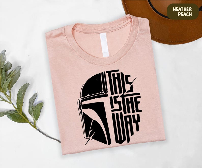 A pink t-shirt with a graphic design featuring a helmet and the text "THIS IS THE WAY" is displayed on a white surface, along with a brown hat and some greenery.