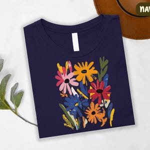 a navy blue t-shirt with a colorful floral design, including yellow, pink, blue, and orange flowers, along with green leaves. The t-shirt is placed on a white surface, and there is a brown hat visible in the background.
