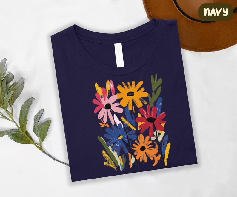 a navy blue t-shirt with a colorful floral design, including yellow, pink, blue, and orange flowers, along with green leaves. The t-shirt is placed on a white surface, and there is a brown hat visible in the background.