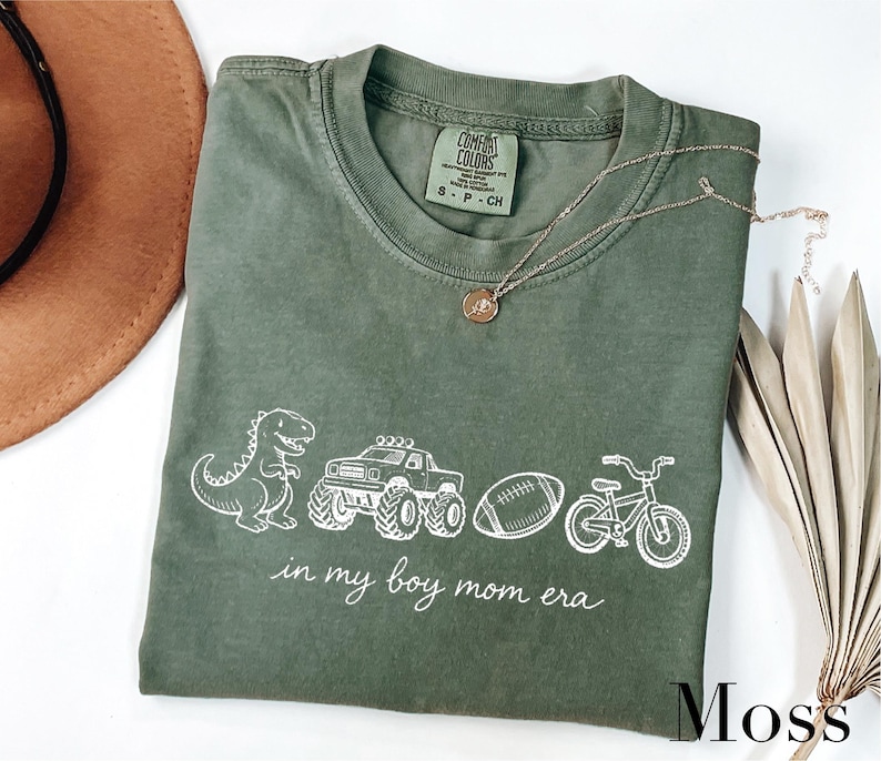A green t-shirt with a graphic design of a dinosaur, truck, bicycle, and football, along with the text "in my boy mom era" printed on it. The t-shirt is laid out on a white surface, accompanied by a brown hat, a necklace, and some dried leaves.